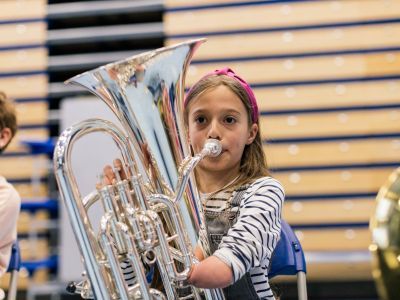 A girl with blonde hair and a pink hair ribbon plays the tuba. She is wearing a striped top and dungarees